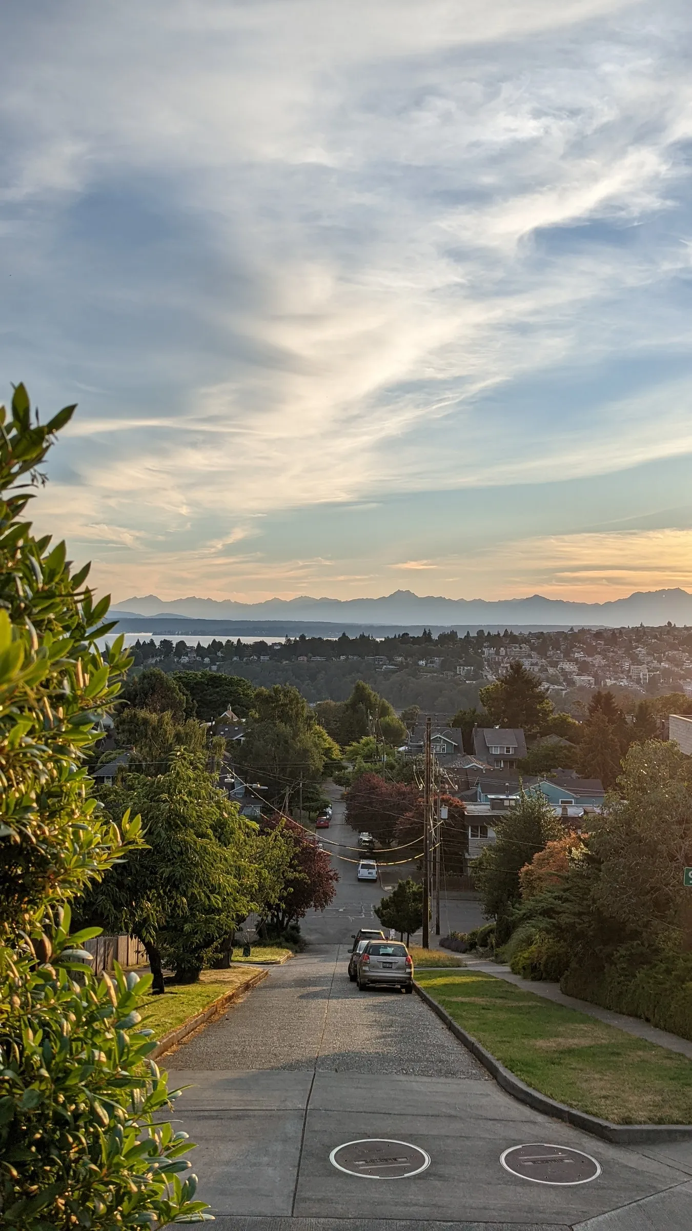 A suburban street descending toward mountains and water under a streaked evening sky.