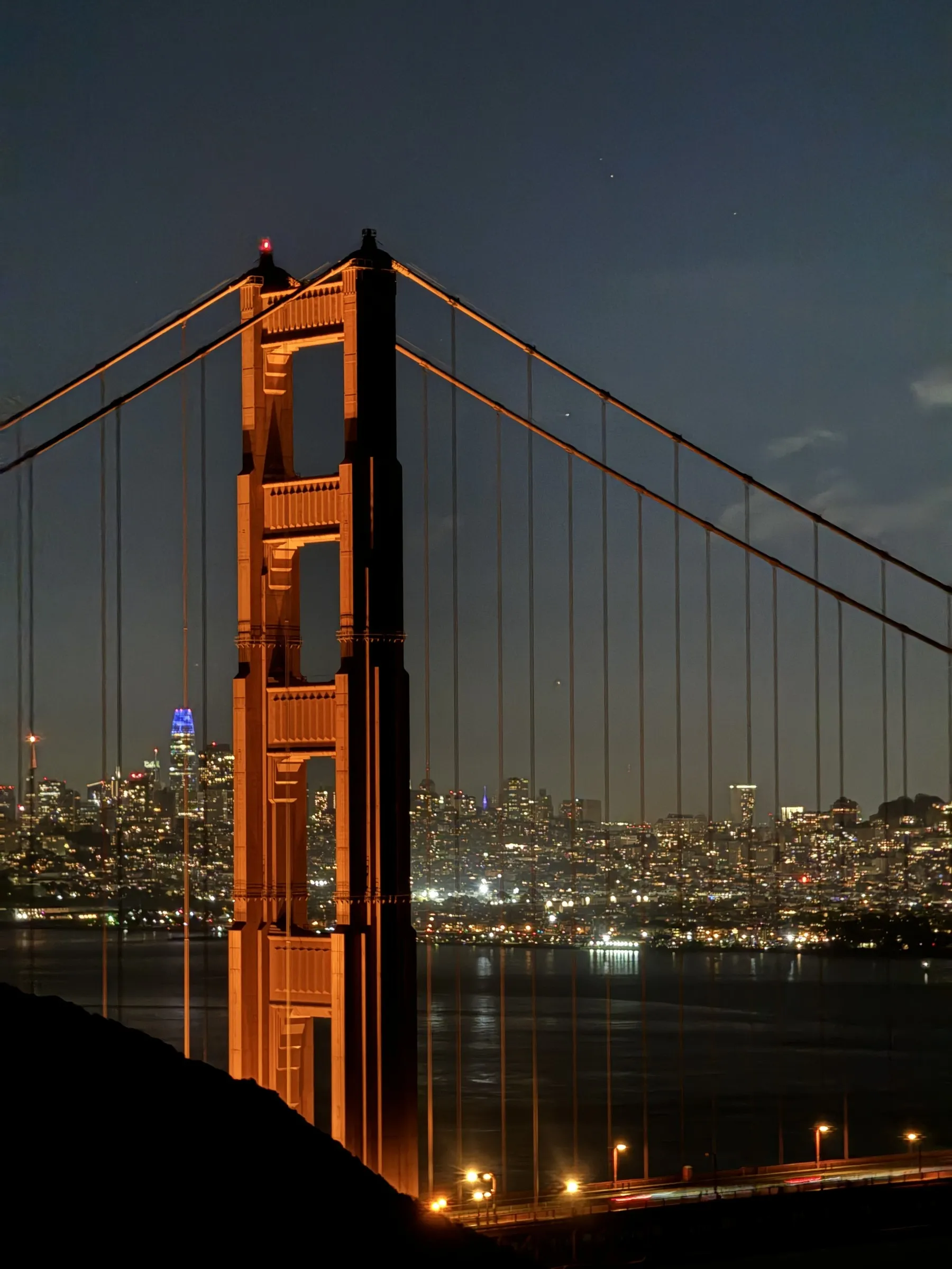 Golden Gate Bridge tower illuminated at twilight with San Francisco city lights in the background.