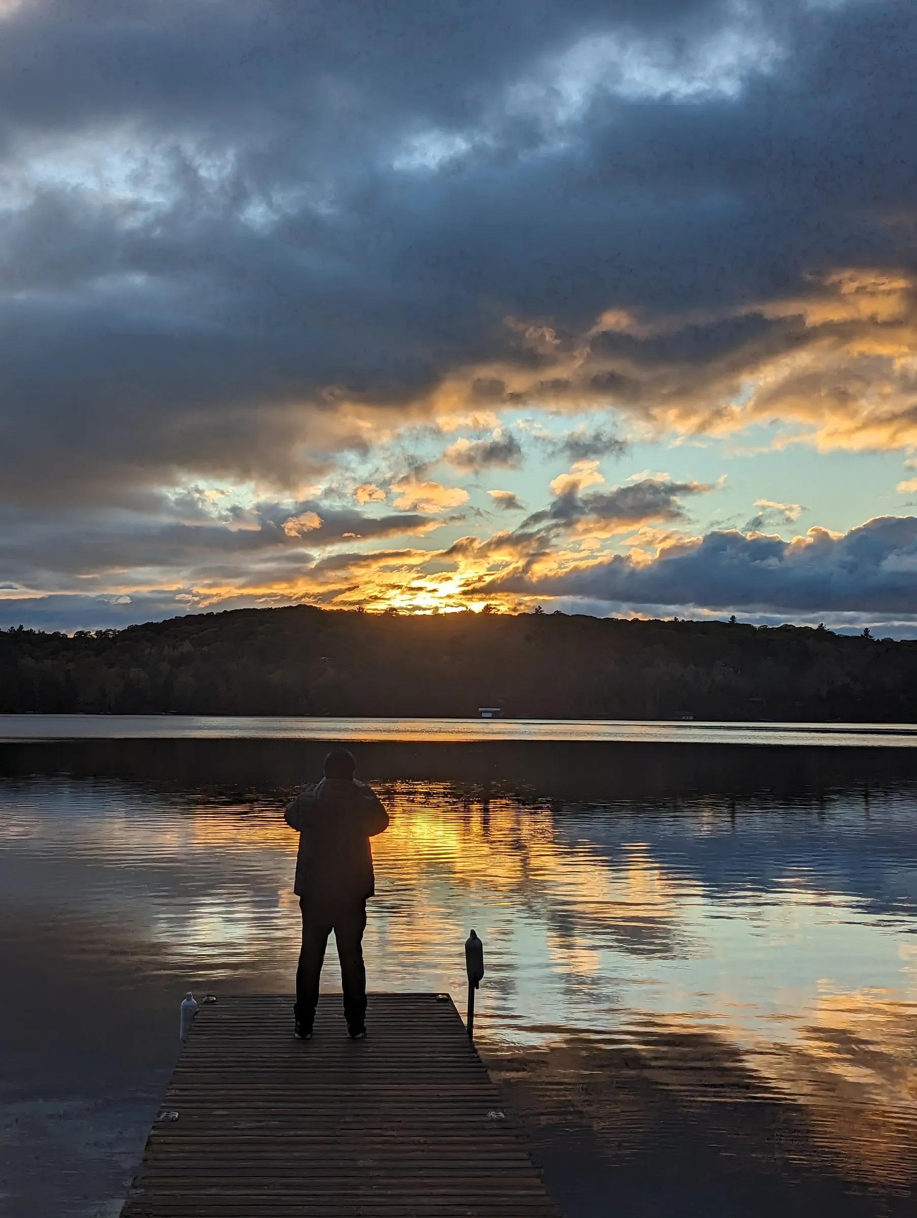 Person standing on a wooden dock facing a dramatic sunset with dark clouds and golden reflections on water.
