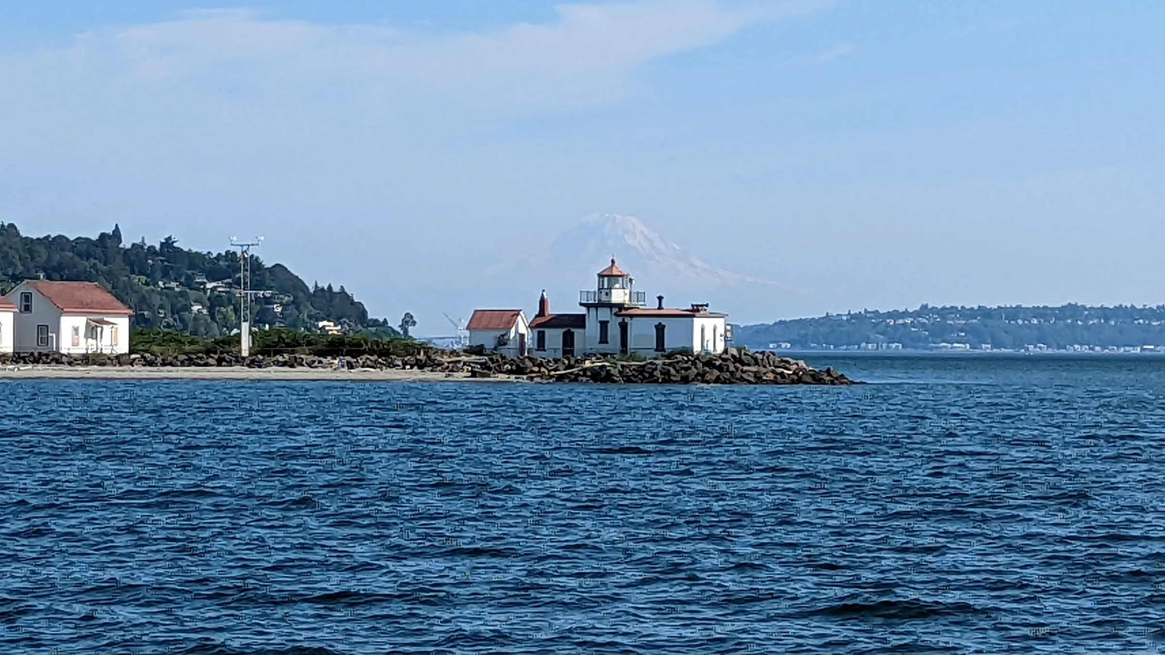 Small rocky island with lighthouse and buildings in blue water with Mount Rainier in background