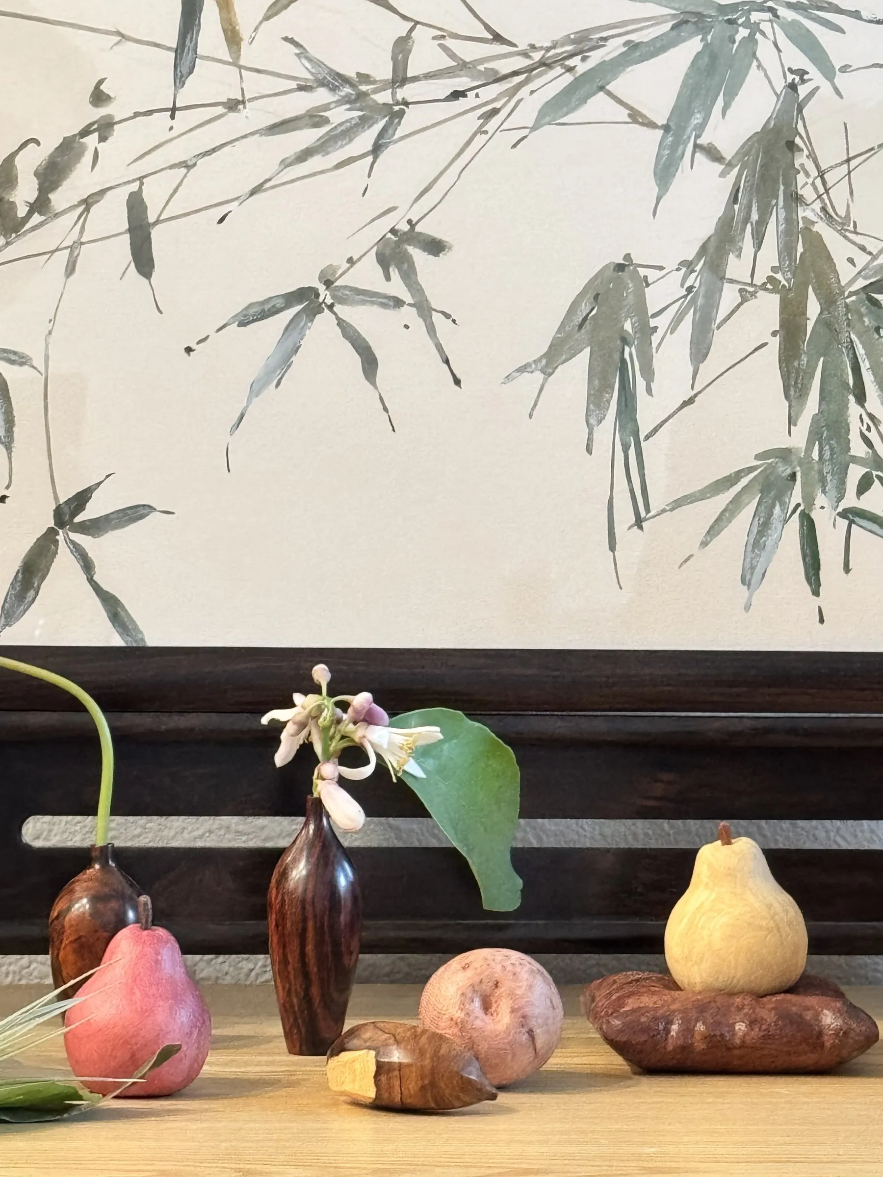 Still life arrangement of ceramic fruit, vase with orchid, and wooden objects on table with bamboo shadow backdrop