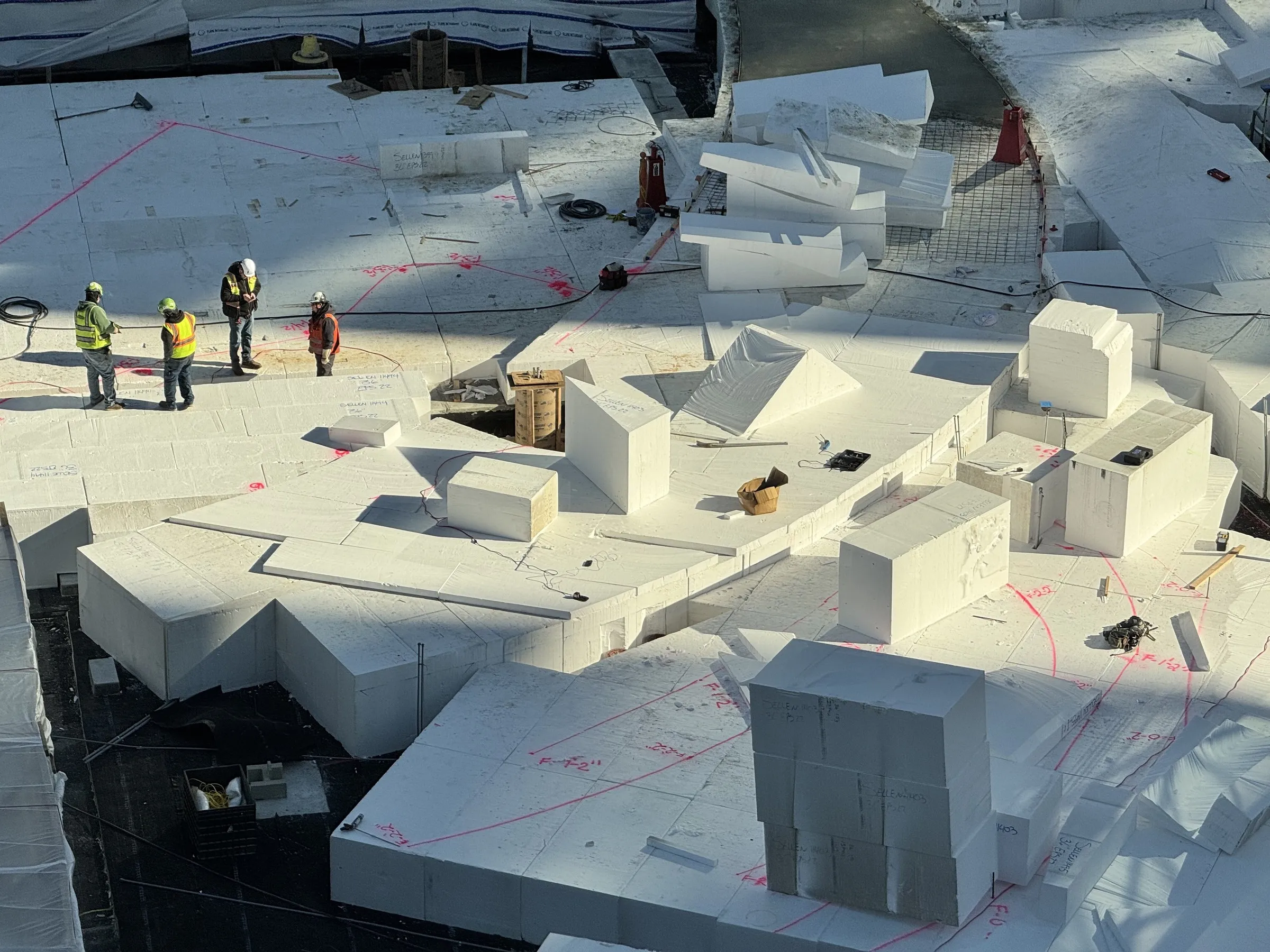 Aerial view of construction workers on white foam-insulated rooftop with geometric blocks and red spray-painted markings.