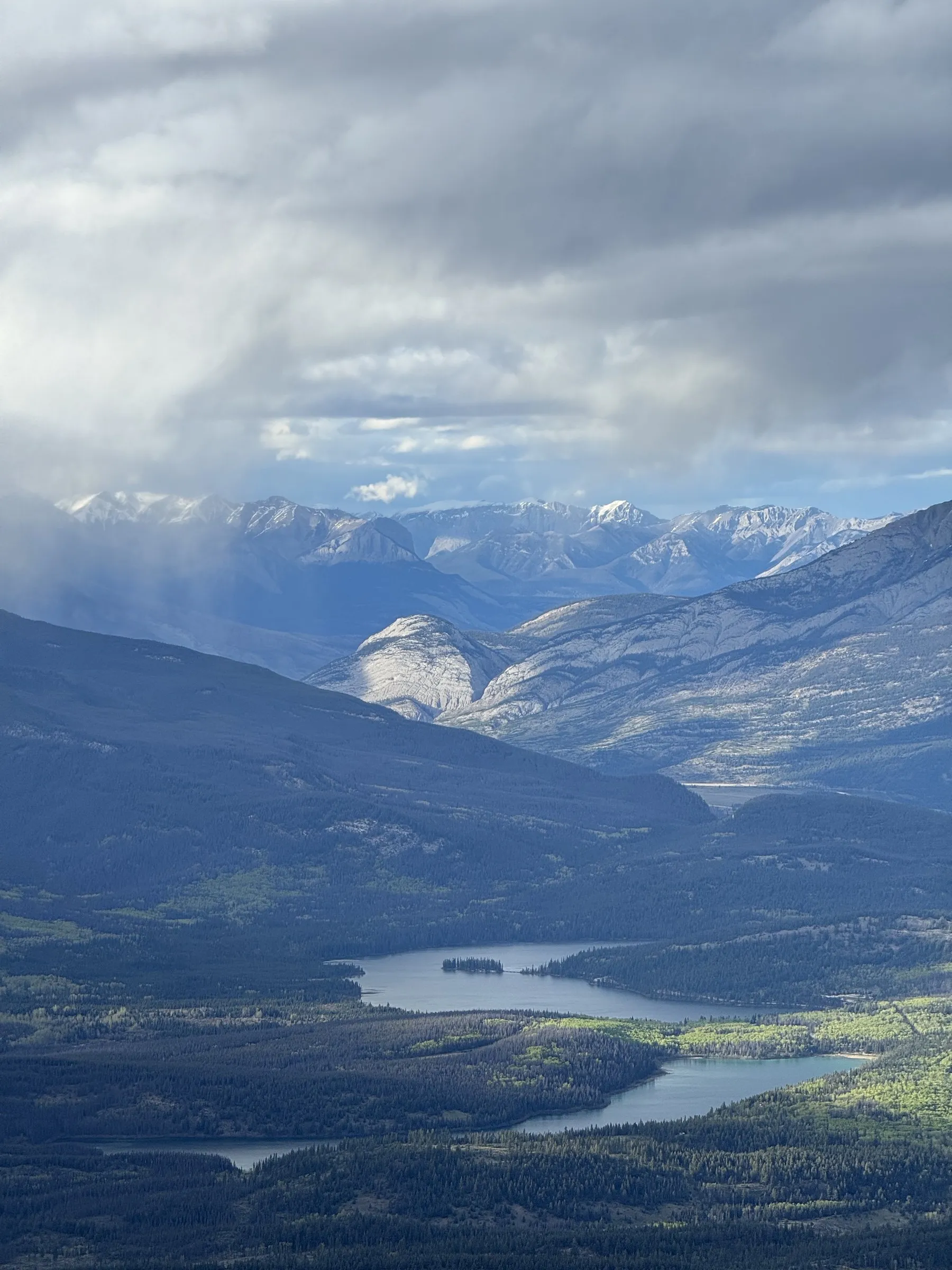 Aerial view of mountain ranges with snow-capped peaks, forested slopes, and two lakes in valley below under cloudy sky.