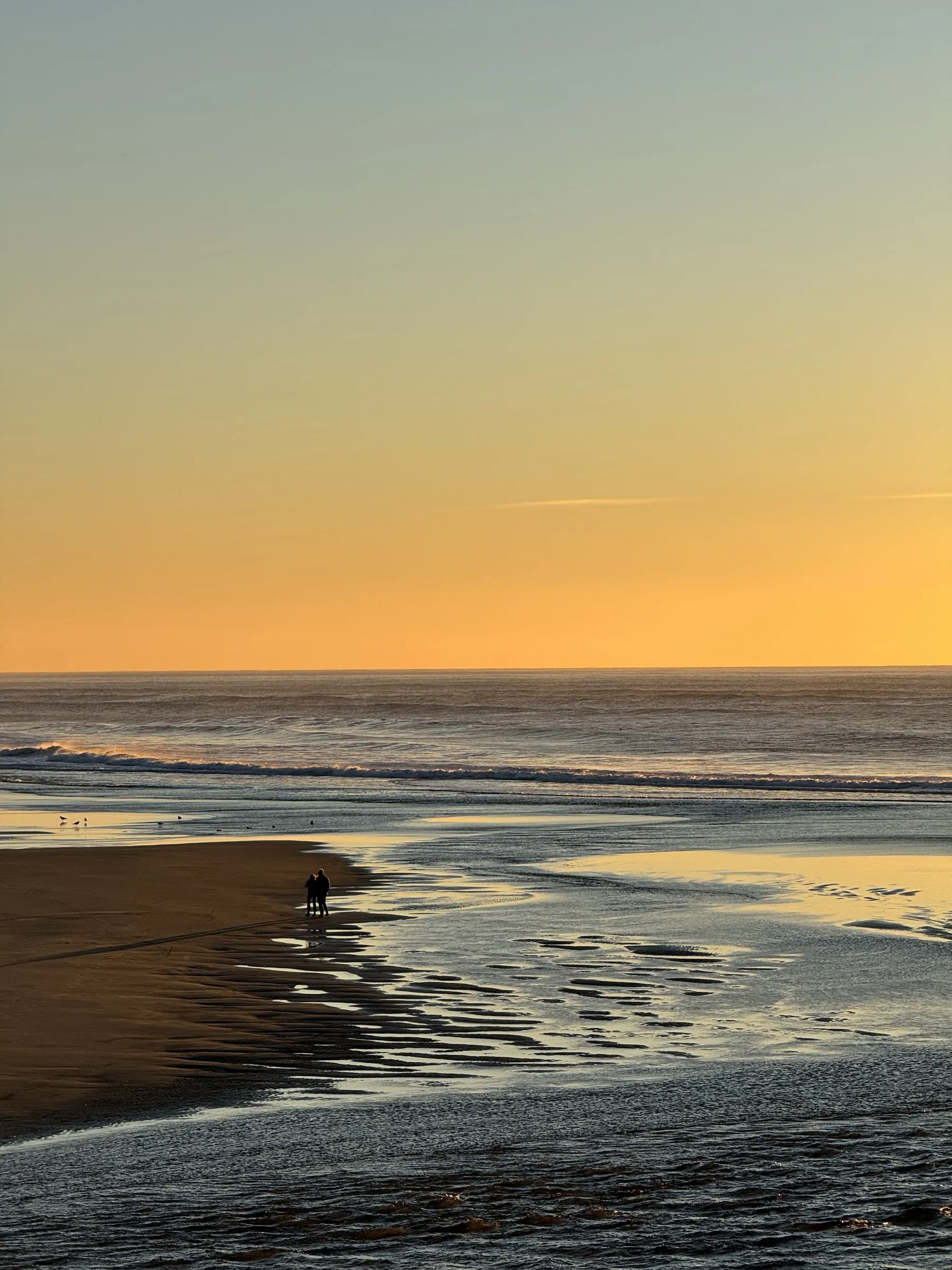 Two people walking on wet beach at sunset with ocean waves and golden sky reflected in sand.