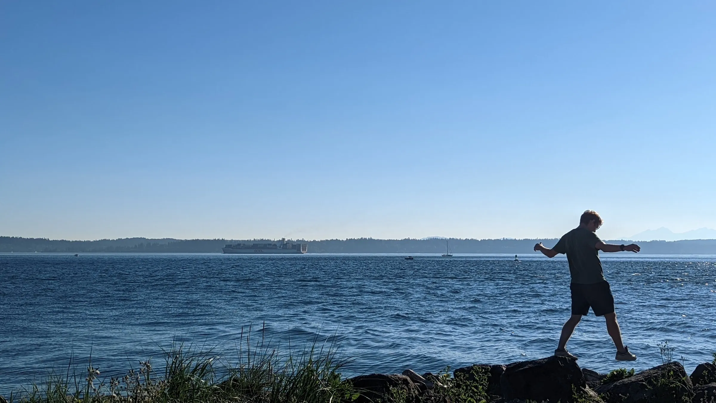 Silhouette of person balancing on rocky shore with arms outstretched against backdrop of calm blue water and distant forested islands.