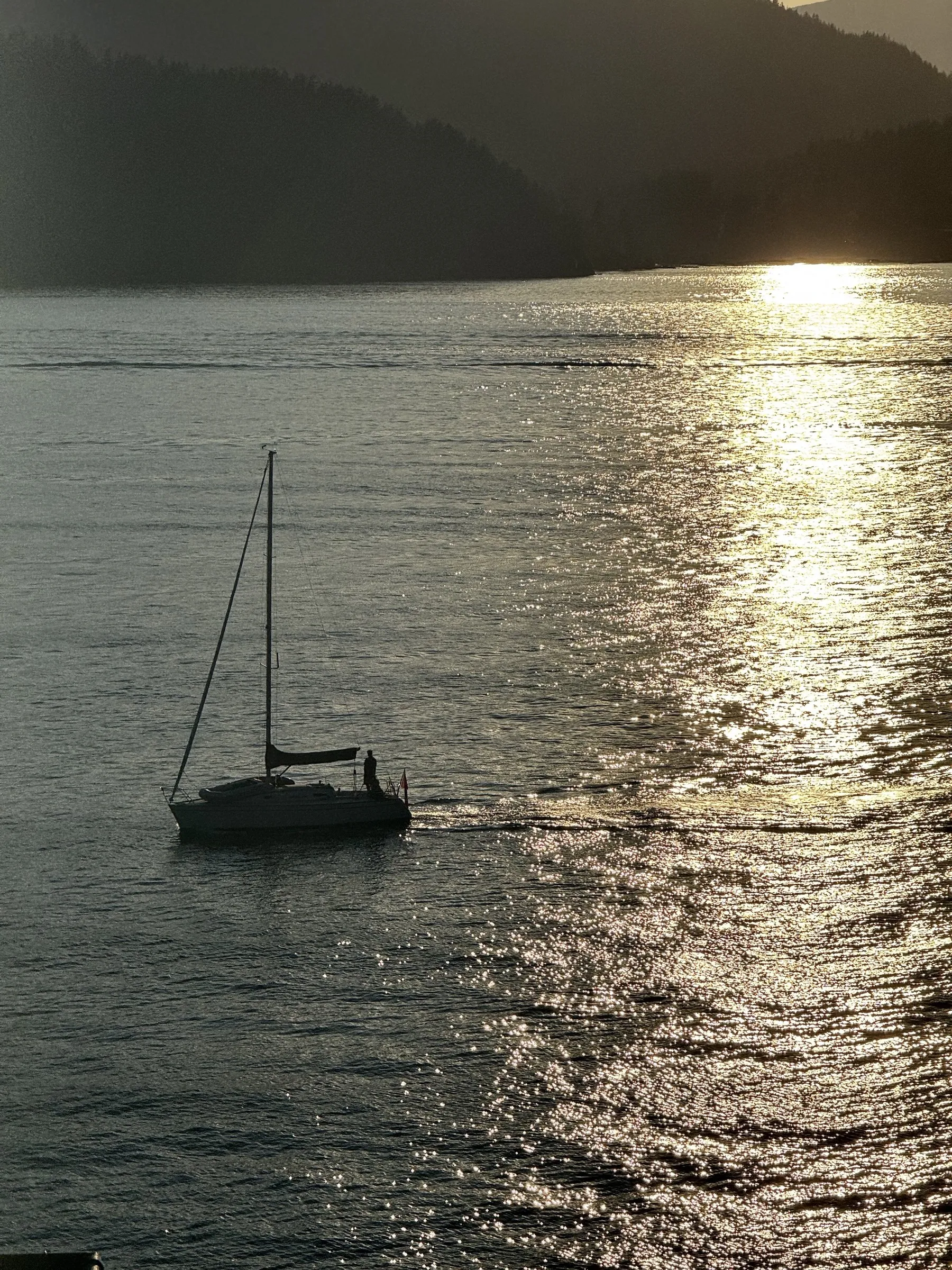 Silhouetted sailboat anchored on calm water at sunset with mountains in background and sun reflecting on water surface.